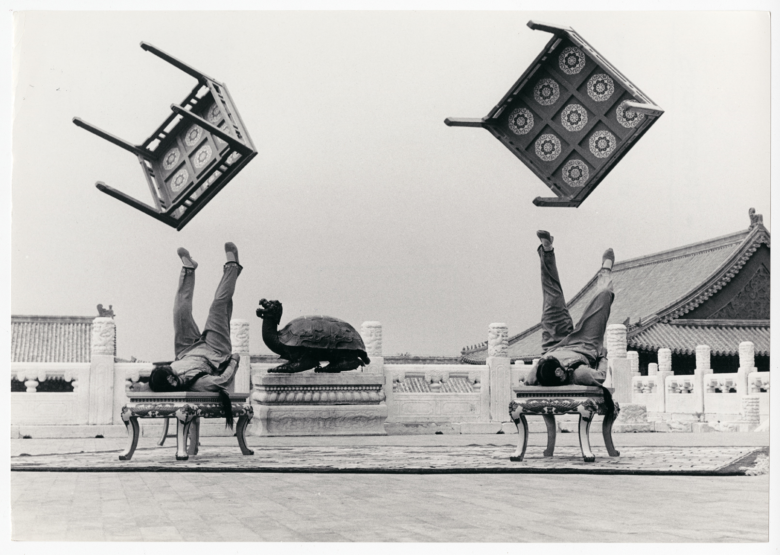 Photograph by unknown artist, black and white image, two gymnasts lie on their backs as two small tables are suspended in the air, as they poise to catch them again