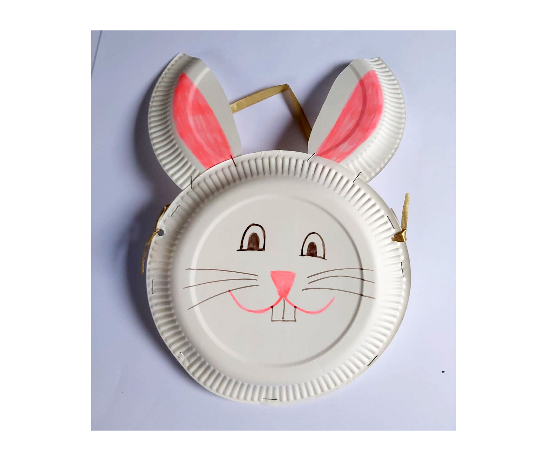 Paper plate lying flat on a table and decorated with an Easter bunny face in black and pink colours with two bunny ears attached in paper plate materials. A hole punch on each side ties a gold string to create a bag