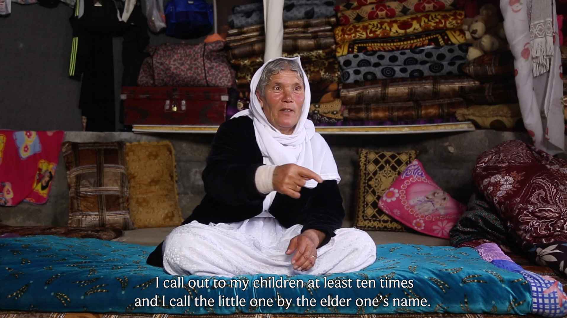 I call out to my children at least ten times and I call the little one by the elder one's name. an elderly woman with short hair and a white veil seated. behind it are many carpets and suitcases