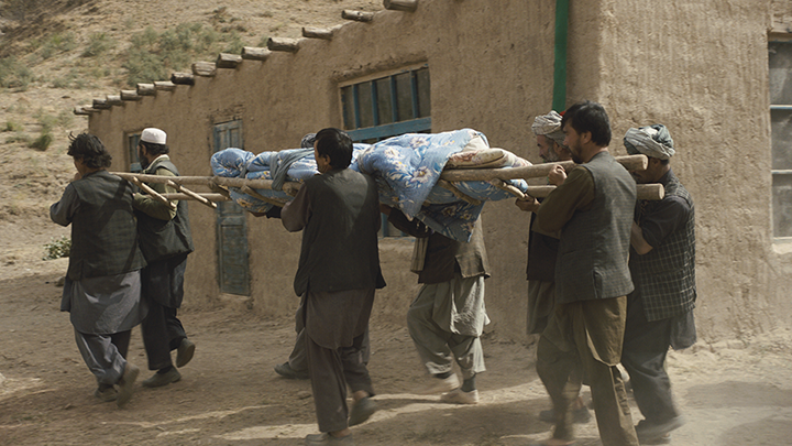 a group of men carry on their shoulders a stretcher with a body wrapped in a blue sheet. in the background there is a house