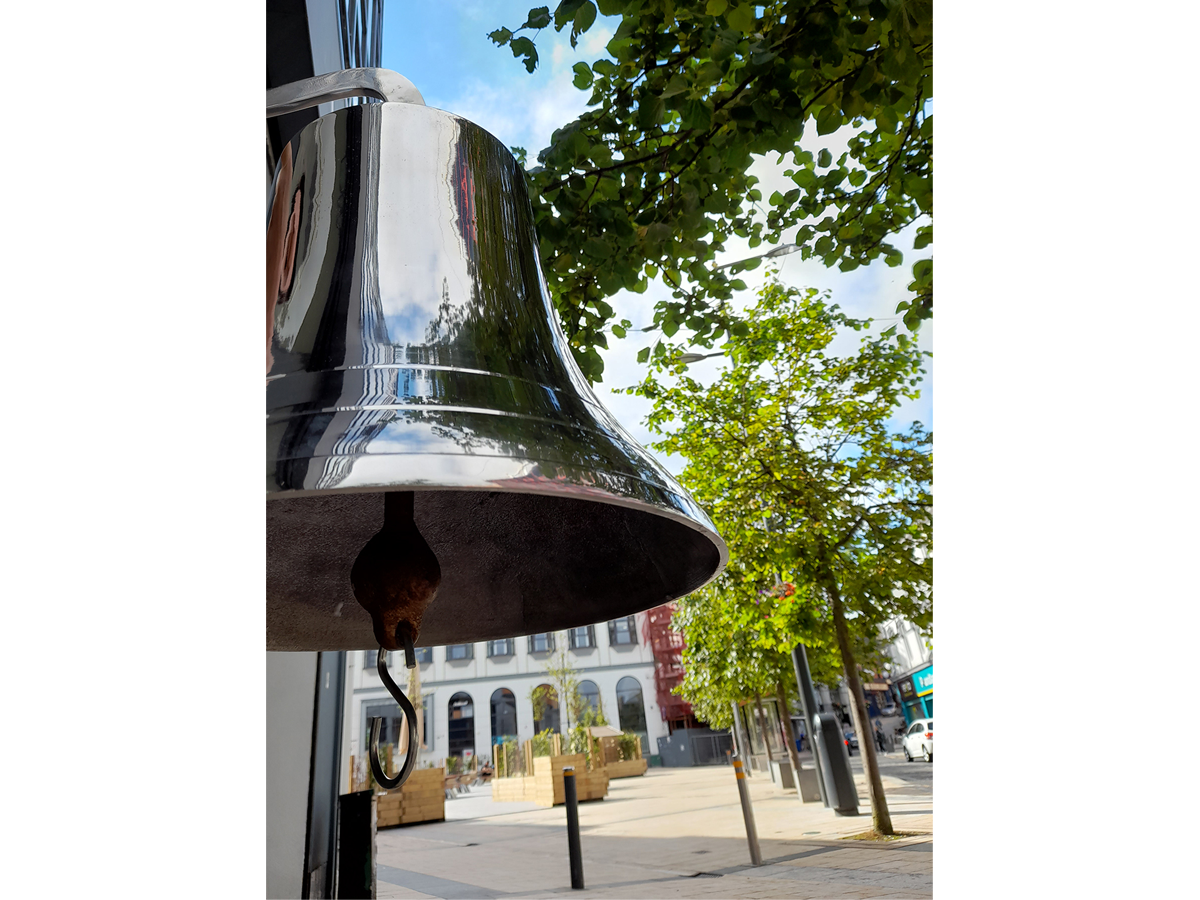 in the foreground there is a bell that reflects trees that are in the background. in the background there is the blue sky and a small square