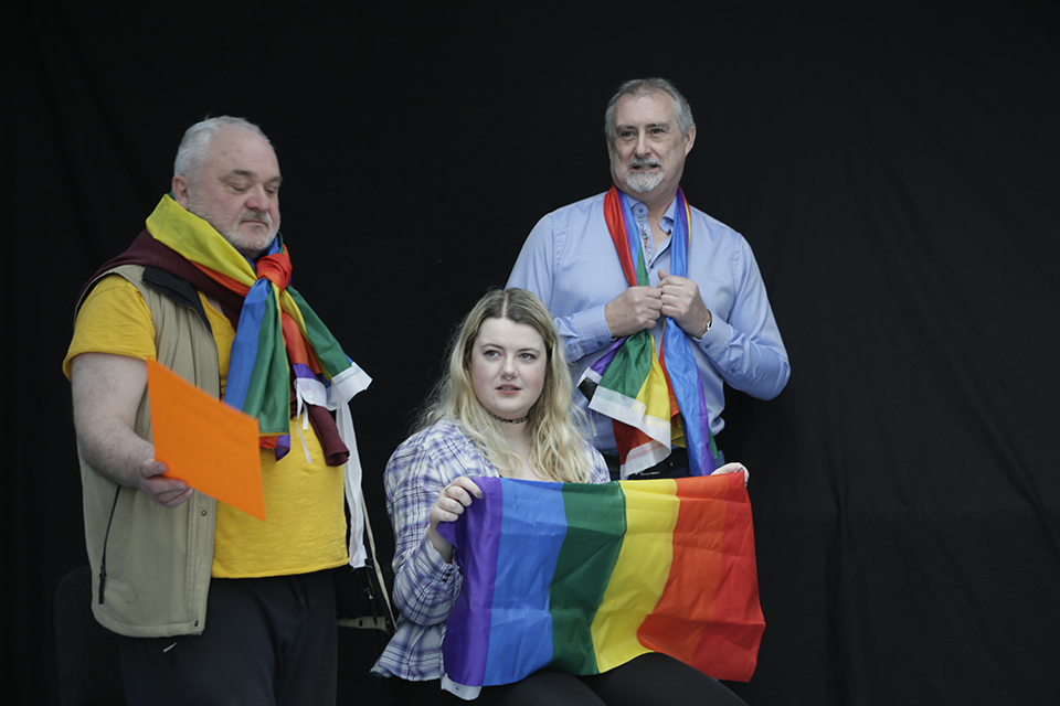 there is a girl sitting in the center with a rainbow flag in her hand, to the left and right two men with a flag each. behind a black background