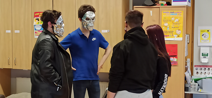 four people talking in a room. two have a silver mask