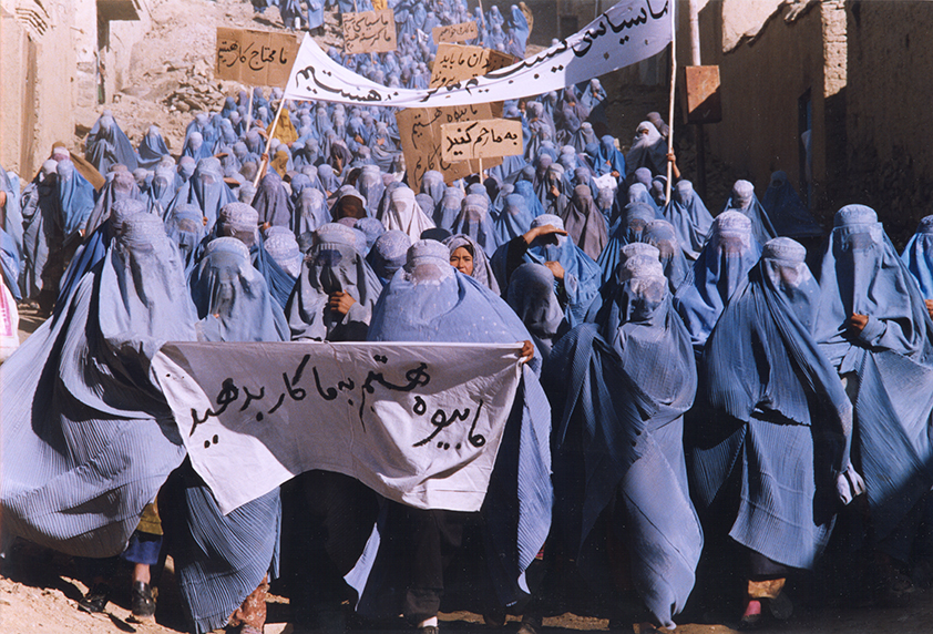 a procession of a protest. people are dressed in blue clothes and have white banners. the buildings around are brown