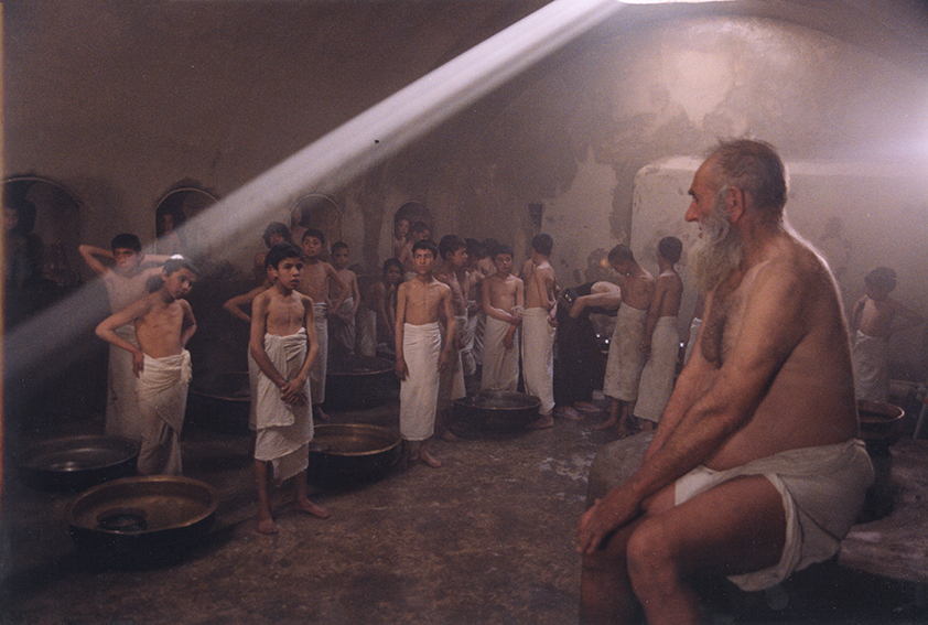 a group of children watching an elderly gentleman. they are dressed in towels, they are in a gloomy room