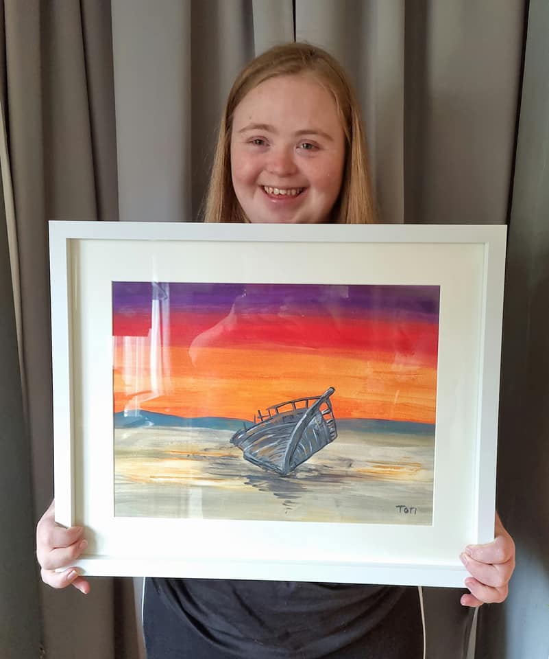 Teenage girl holds up brightly coloured painting depicting a whale's fin entering the water at sunset.