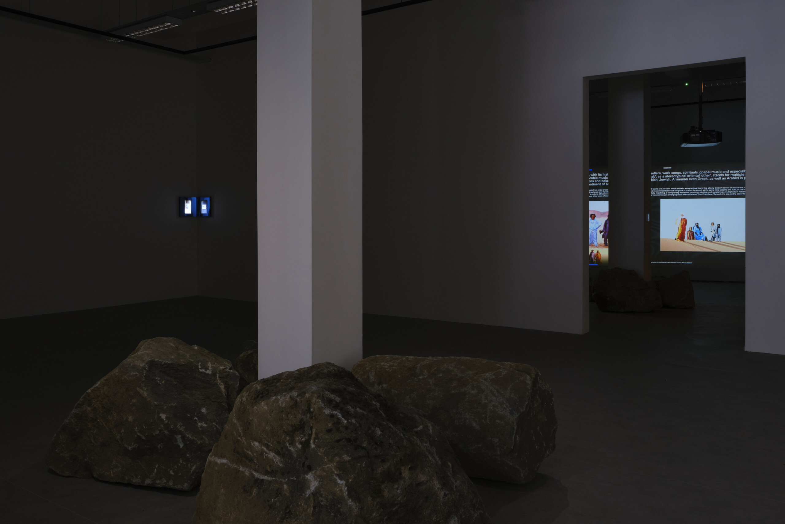 view of two rooms of an exhibition. in the first there is a column with stones on the floor. in the second there is a projected text