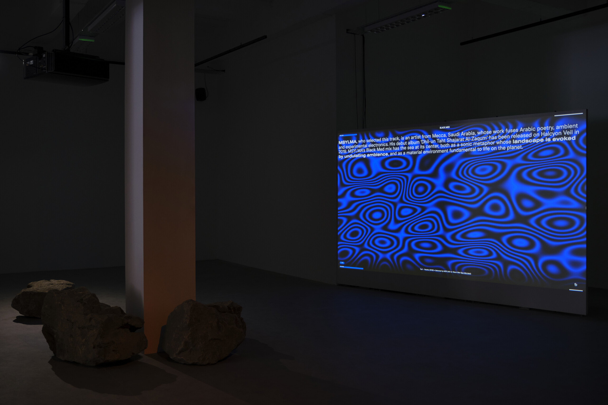 dark room with a column to the left, with stones at its feet. To the right is a large TV