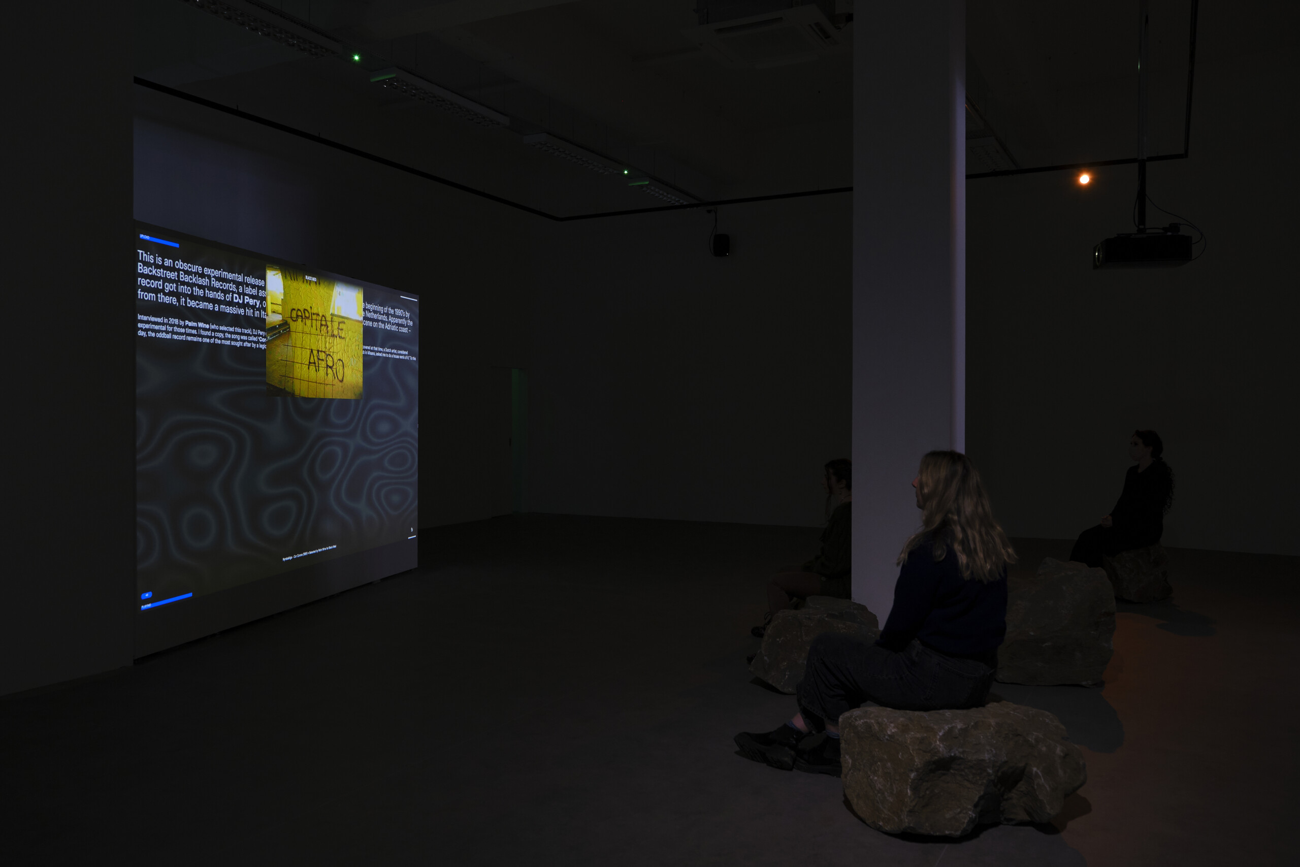 two girls sitting on stones right at the foot of a white column. To the right is a projected work