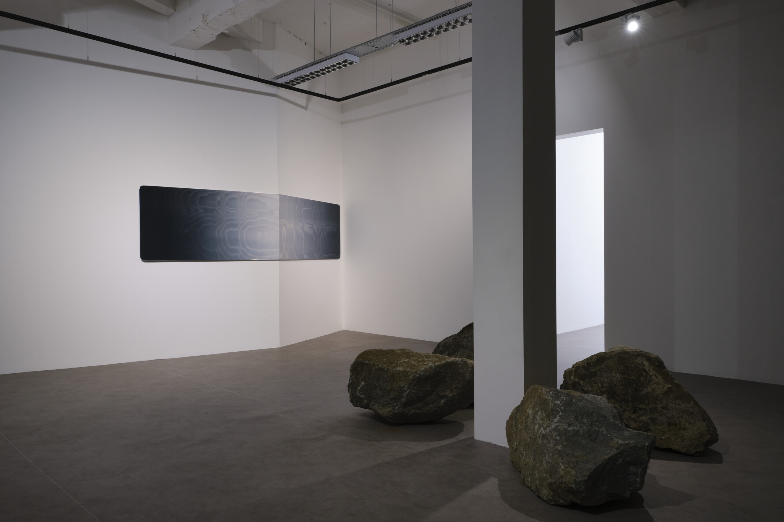 white room with a work of art on the left wall. to the right is a column with stones on the floor