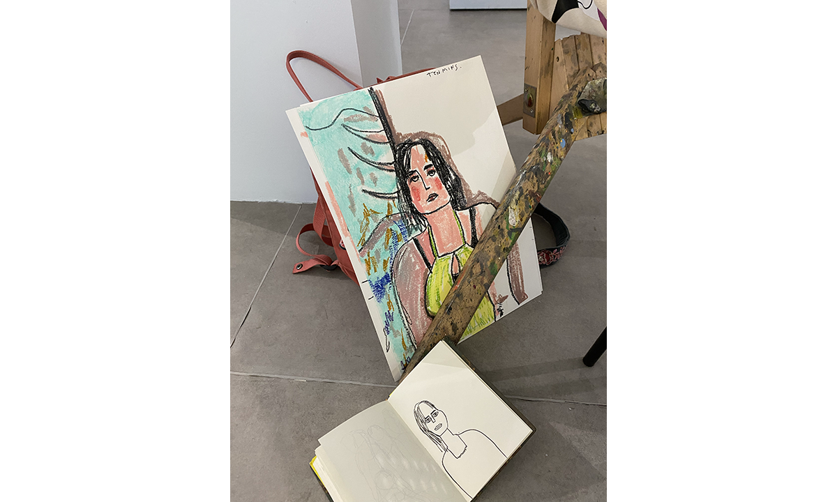 Cement floor with art sketchpad and painting sitting facing viewer. Coloured drawing of a woman with brown hair and yellow top.