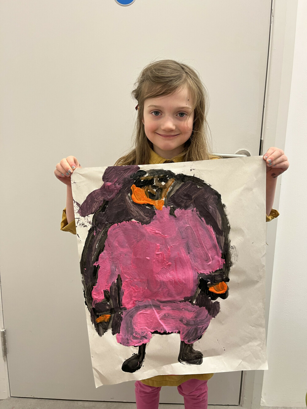 Female child with long brown hair and a fringe stands in front of a white wall holding a large sheet with a painting on it, in colours black, pink and orange. She smiles at the camera.