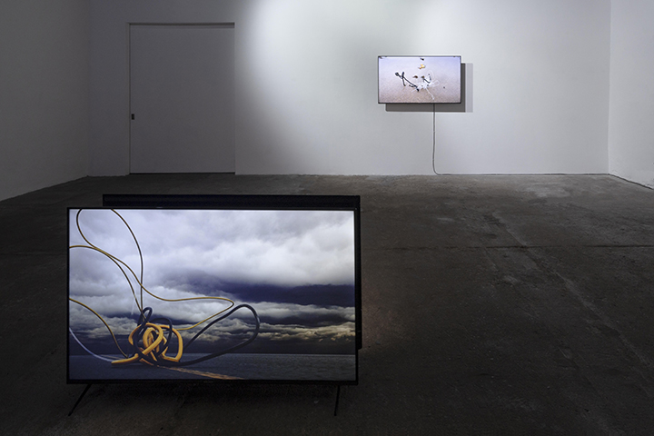 Cement grey floor with white wall running around the background of the image. A TV screen sits facing the viewer on the floor and depicts a grey cloudy sky with a bundle of yellow and black wires. A TV screen is shown hanging on the wall in the background