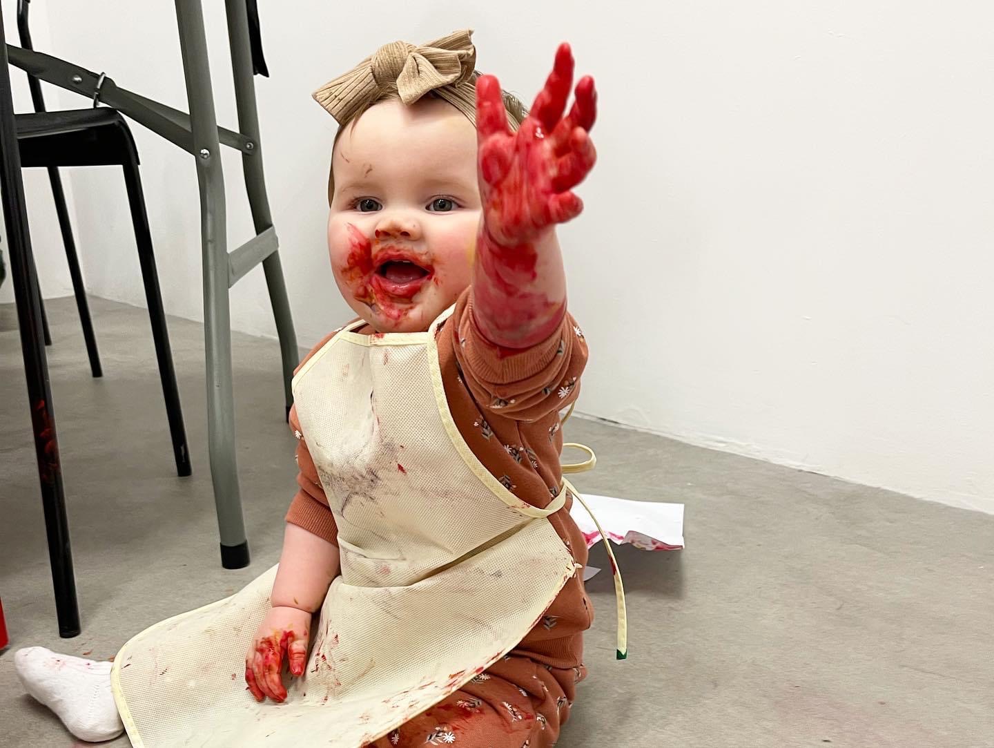 1 year old child sits on the floor with a beige apron on and holds her left hand up towards someone she is communicating with in the background. Her hand and mouth are covered in red paint.