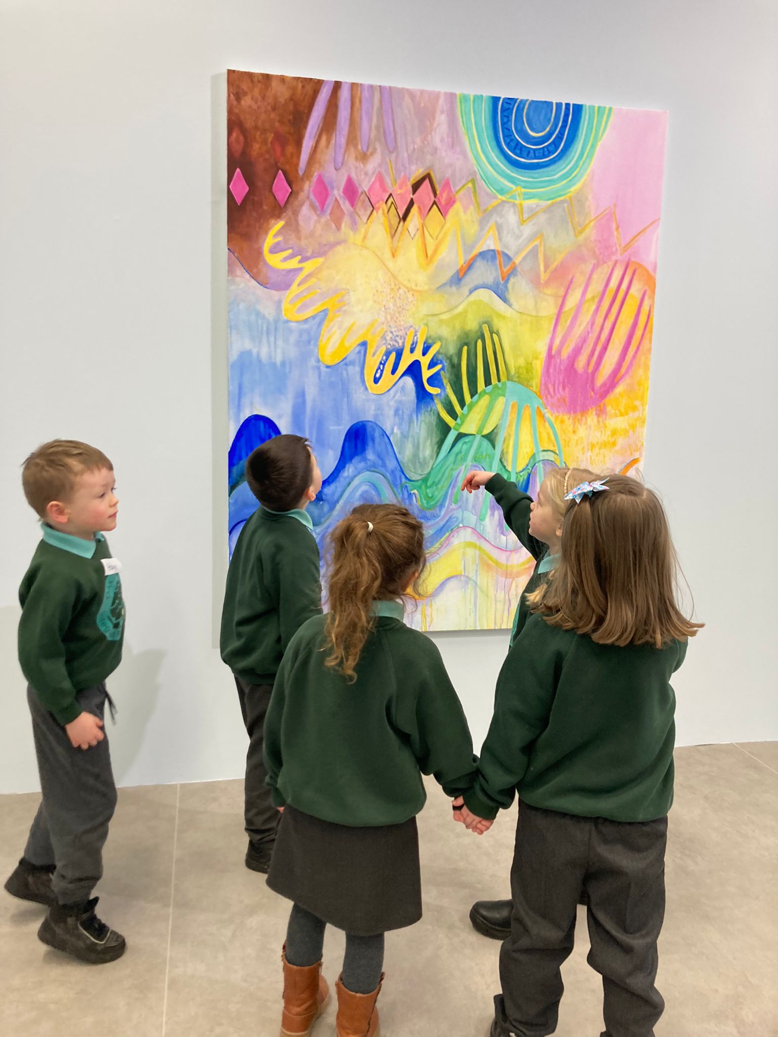 Four children stand facing away from the viewer, looking and pointing at a colourful painting hanging on a white wall. They are wearing school uniforms.