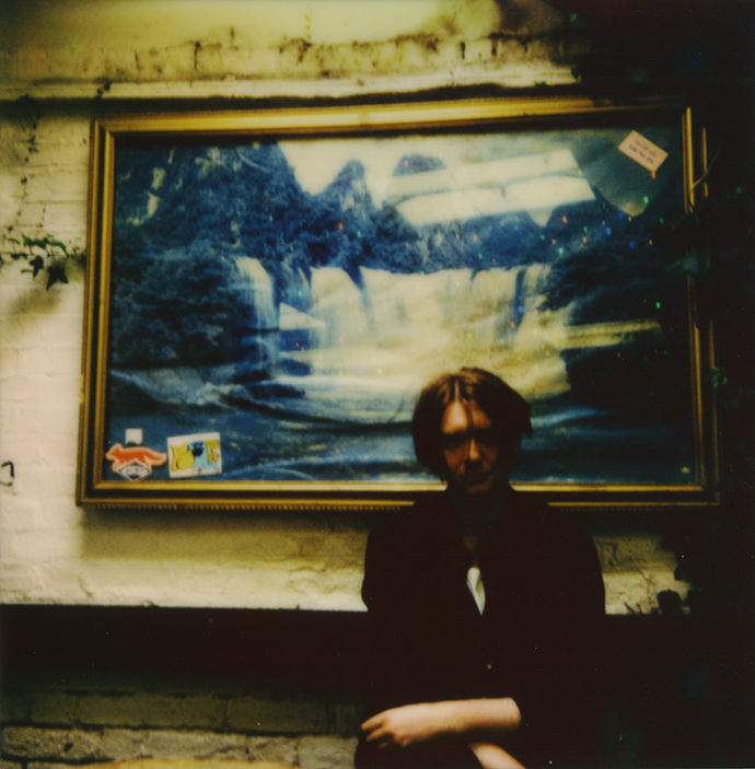 Blurred image of a young man wearing a black coat with medium length auburn hair standing in front of a wall with a large gold framed painting behind him.