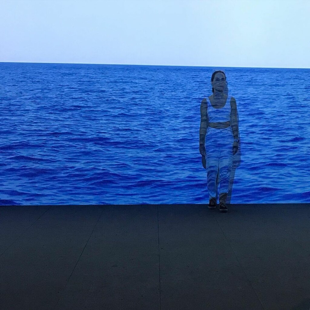 a woman standing in front of a wall onto which the sea is projected.