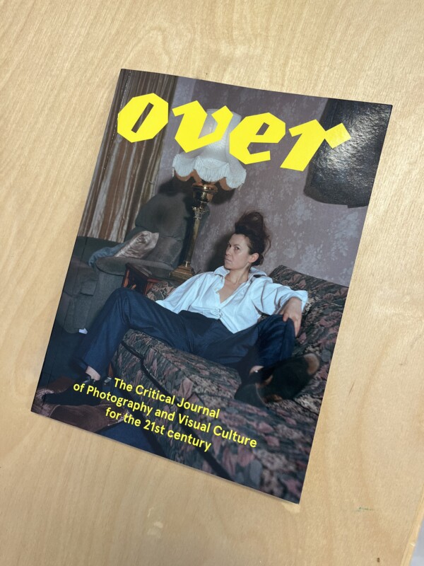 The image of a publication cover with the title 'Over' at the top in yellow. It shows a woman sitting on a couch with a stern look on her face.