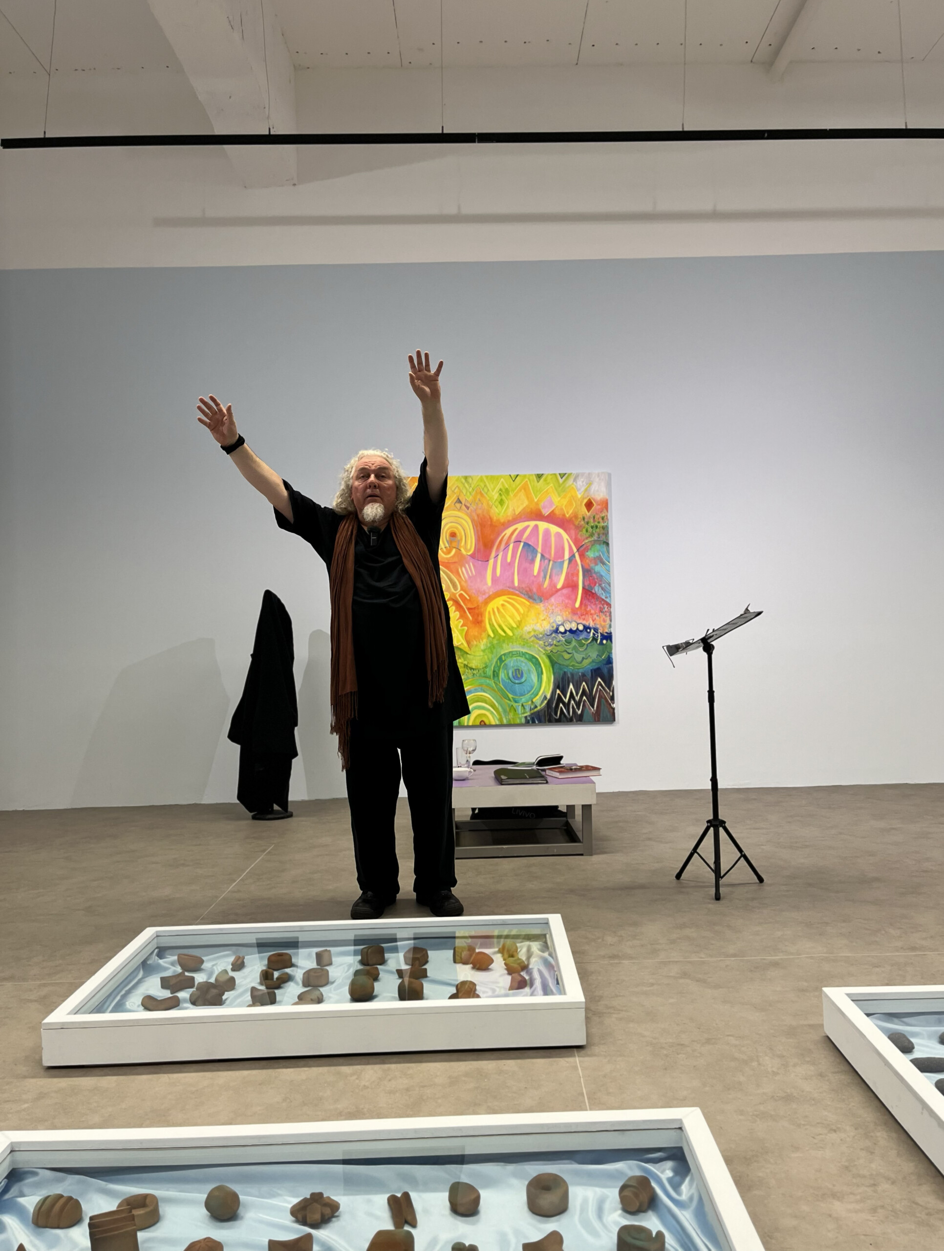 Man with long white hair stands with his arms raised above his head. He in in a large room with blue walls and a colourful image on the wall behind him. There are vitrines on the ground with dark objects inside.